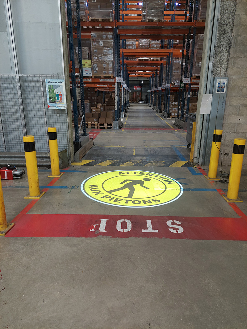 Projection lumineuse jaune au sol d'un panneau de s&eacute;curit&eacute; "Attention aux Pi&eacute;tons" &agrave; l'entr&eacute;e d'une all&eacute;e d'entrep&ocirc;t logistique.
