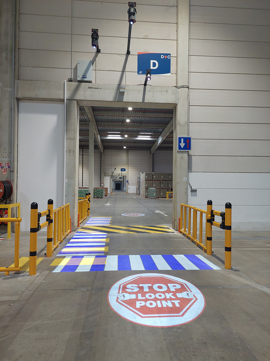 Passage pi&eacute;ton virtuel bleu et blanc et pictogramme "STOP LOOK POINT" projet&eacute;s au sol &agrave; l'entr&eacute;e d'une zone logistique.